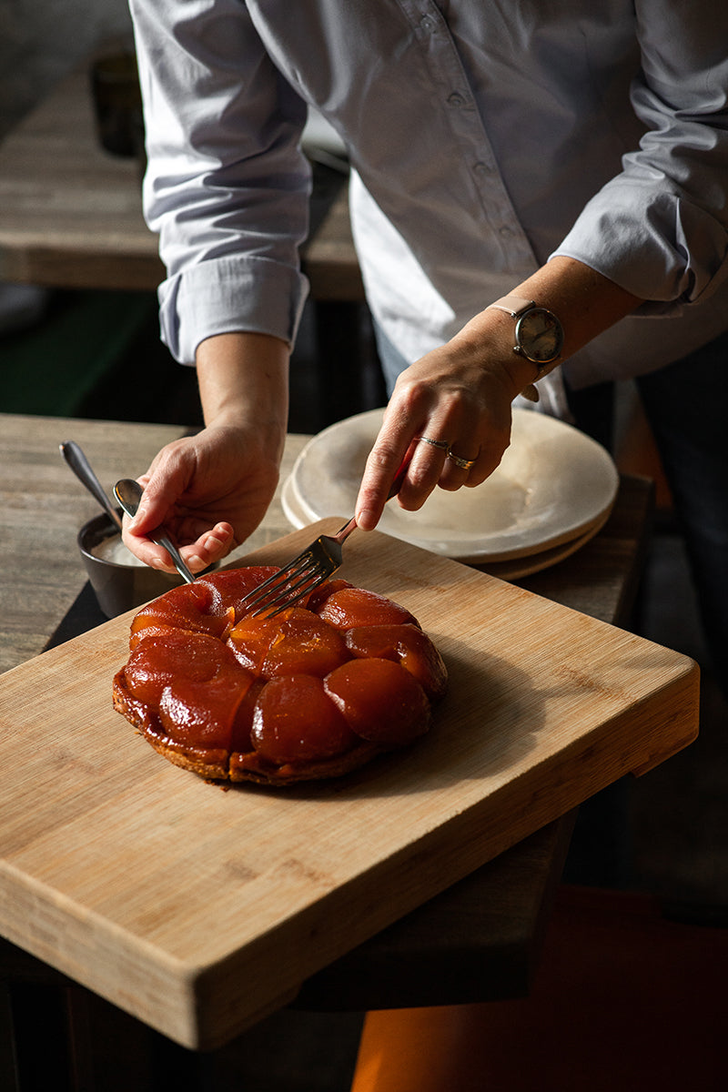 Une personne coupe un dessert caramélisé en forme de tarte sur une planche en bois. Elle utilise une fourchette et un couteau pour servir.
