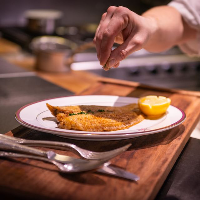 Chef ajoutant une touche finale à un filet de poisson doré servi avec un quartier de citron.