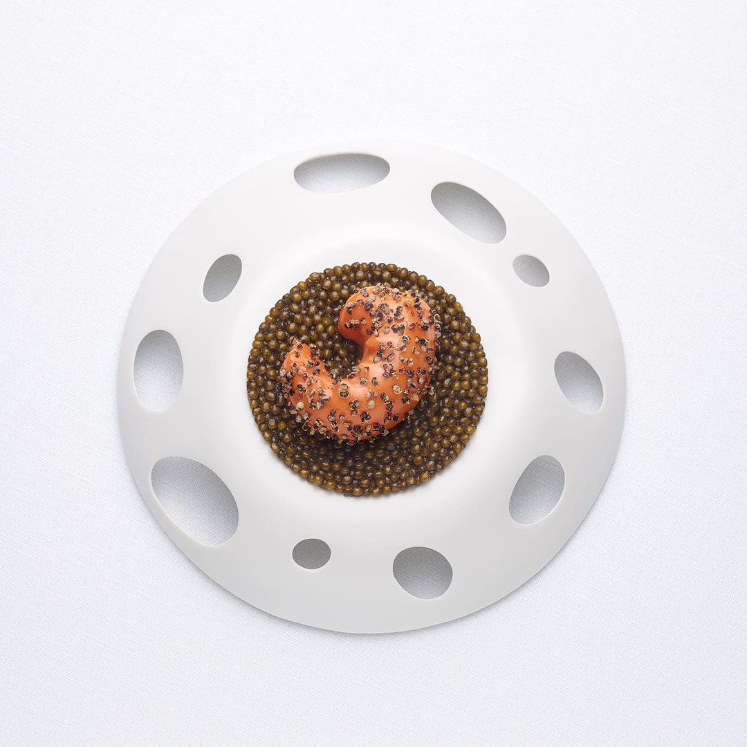 Un plat gastronomique composé d'un morceau de saumon en croûte de graines de quinoa, disposé sur un lit de perles de caviar, le tout présenté dans une assiette blanche percée de trous circulaires pour un effet artistique.