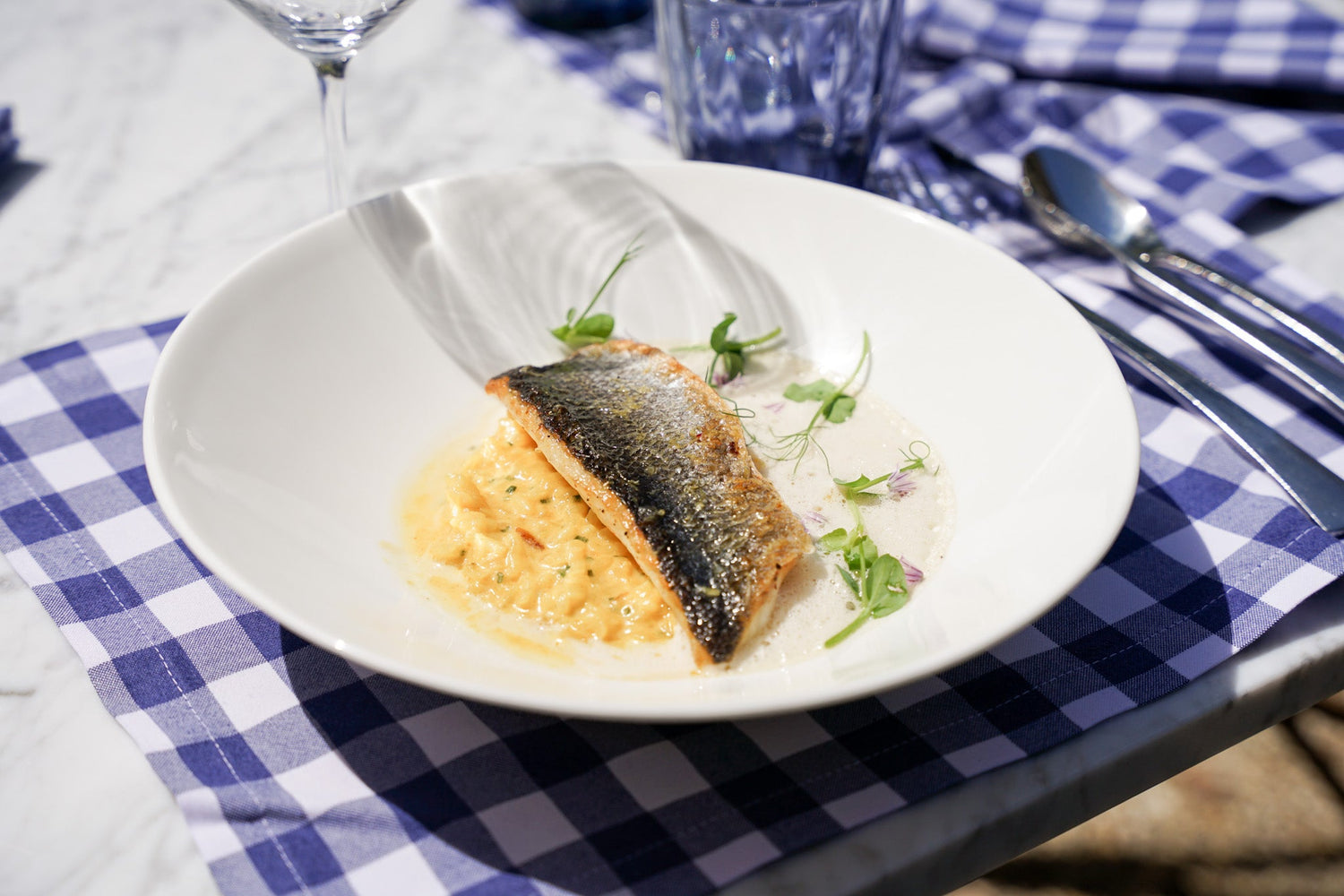 Plat principal avec un morceau de poisson grillé sur un lit de risotto crémeux, garni de micro-pousses, servi dans une assiette blanche avec des serviettes à carreaux bleus.