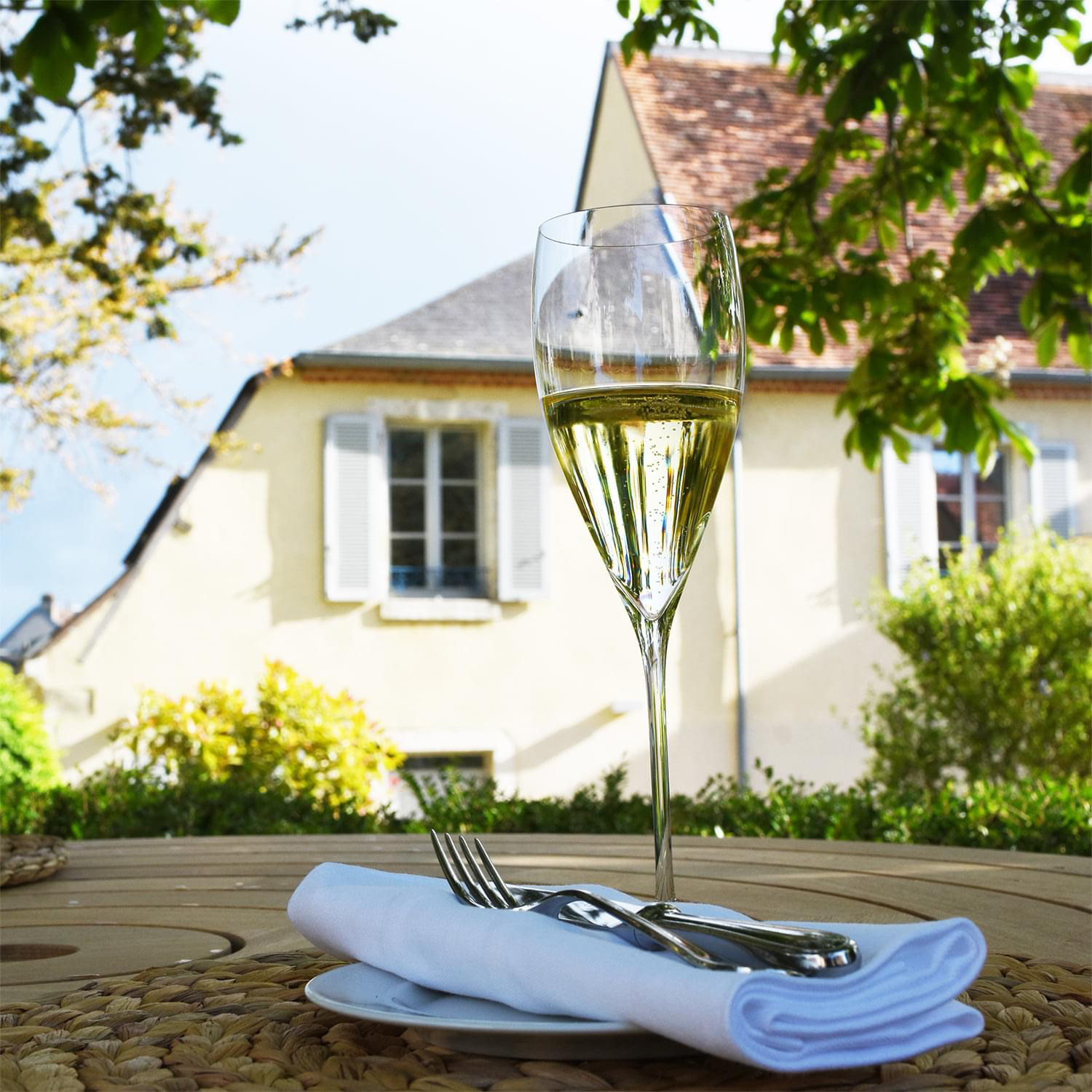 Verre de champagne posé sur une table extérieure avec une serviette et des couverts, avec en arrière-plan la façade d'une maison sous un ciel dégagé.